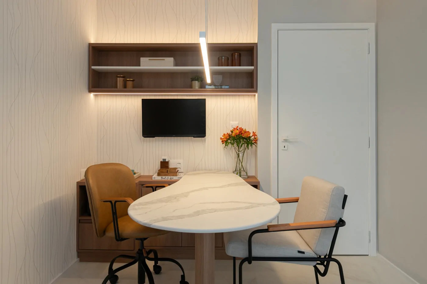 Consultório com mesa oval de mármore branco, cadeiras em couro caramelo, nichos em madeira e parede ripada clara