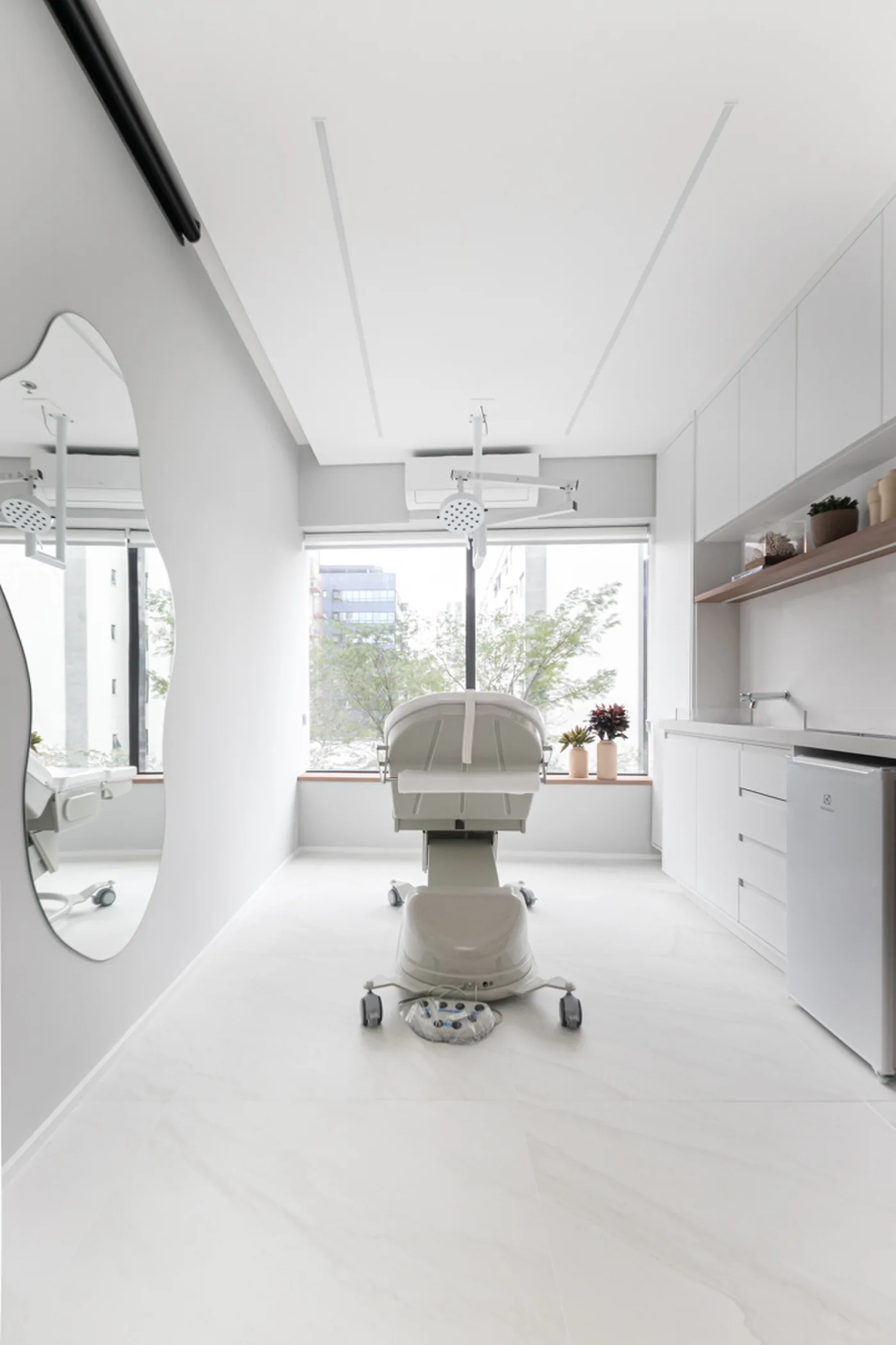 Sala de procedimentos clara em arquitetura clínica médica, com cadeira branca, foco de luz no teto e espelho orgânico