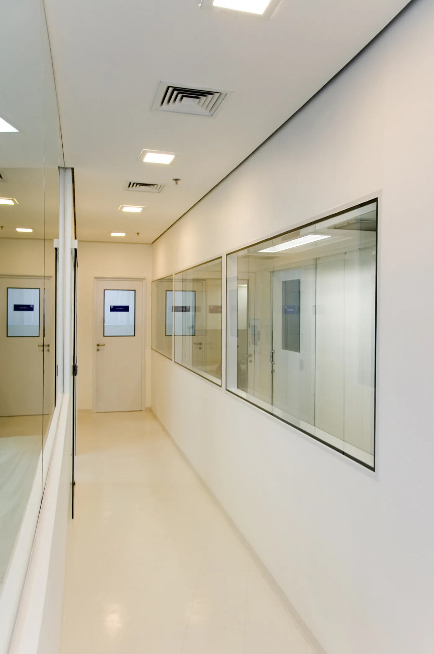 Corredor branco com portas e janelas de vidro internas, piso claro brilhante e forro com luminárias e grelhas de ar